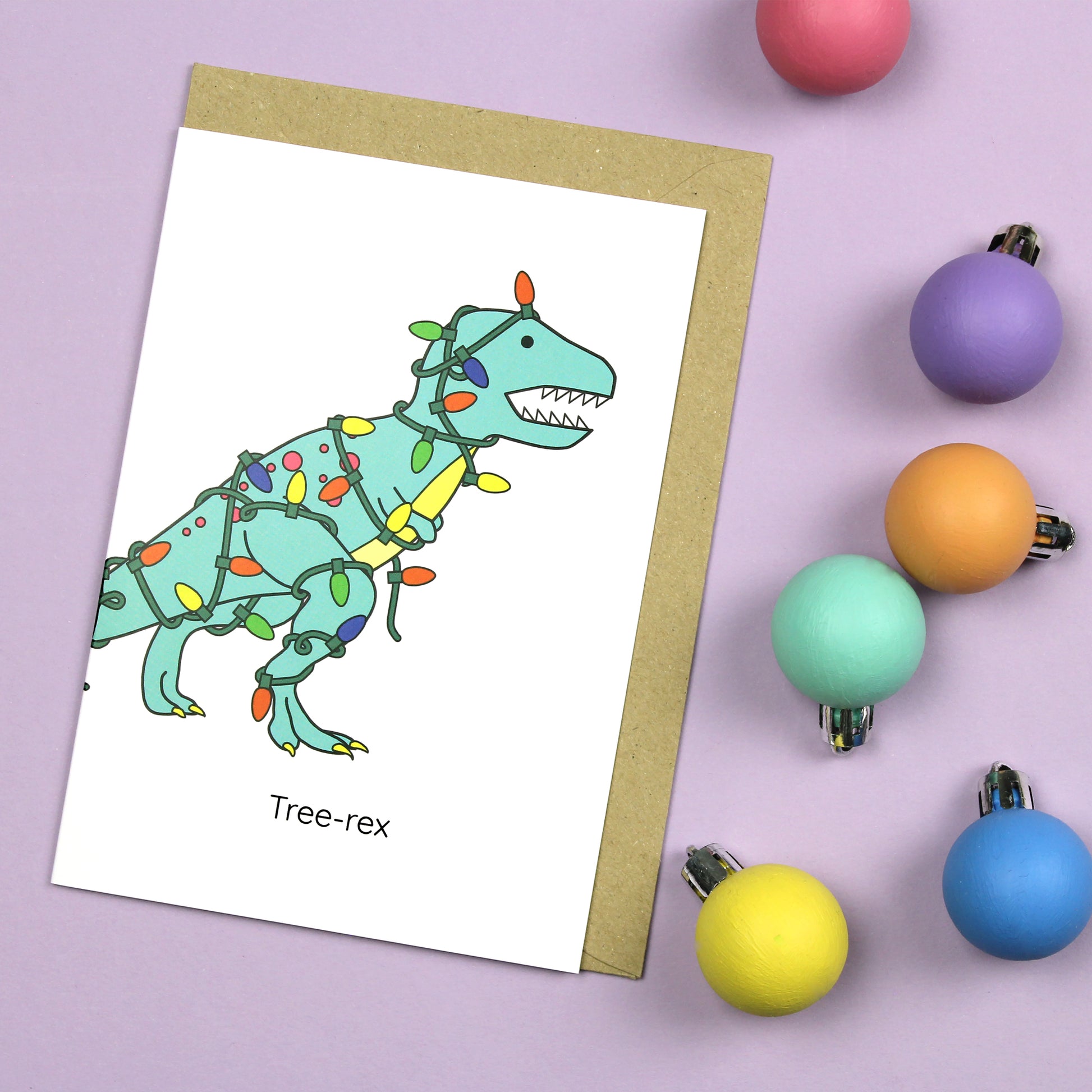Tree-rex greeting card with a brown kraft envelope on a purple background with pastel coloured baubles scattered next to it