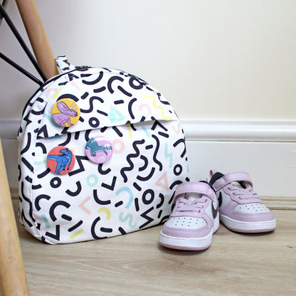 memphis print kids backpack with 3 mini dinosaur badges, next to toddler shoes and a chair