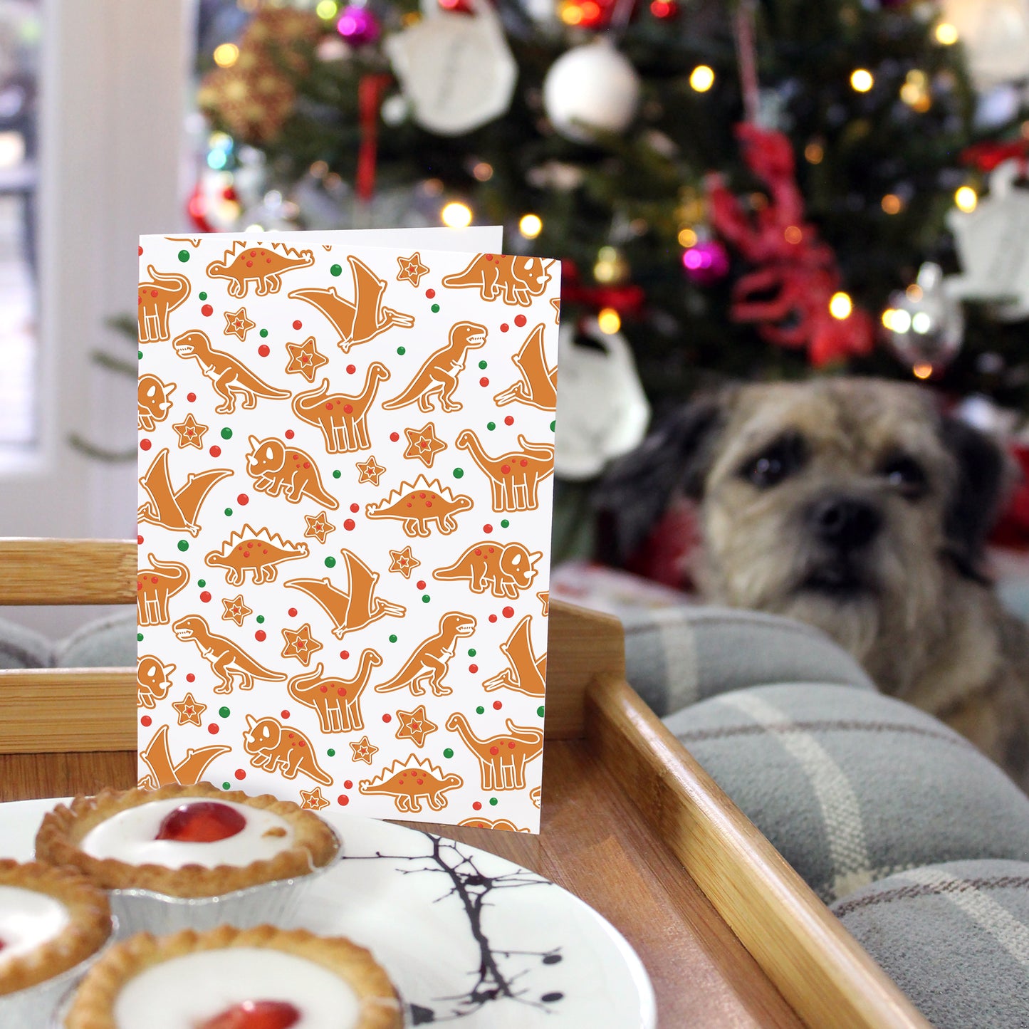 dinosaur gingerbread Christmas card on a tray with a plate of bakewell tarts. in the background is a blurred out Christmas tree and a border terrier dog 