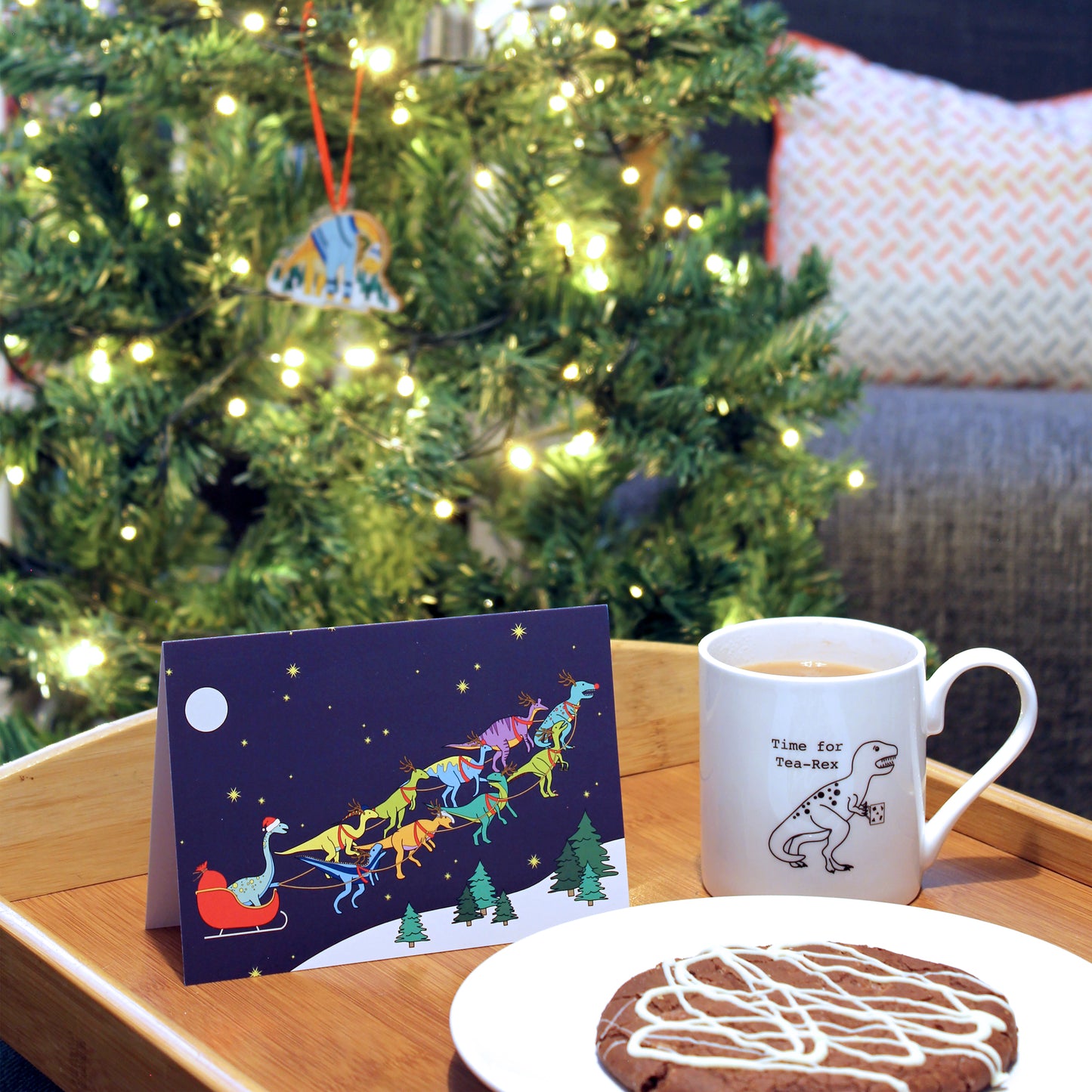 Santa's sleigh dinosaur Christmas card on a tray with tea and a cookie. There is a Christmas tree in the backgorund