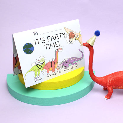 Space Dinosaur Invitation folded on a yellow and a green block of wood with a pink long neck dinosaur figurine wearing a party hat.