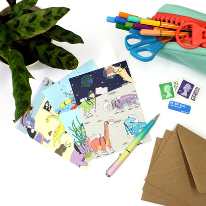 3 dinosaur postcards on a white desk with envelopes, a plant, stamps and a pencil case