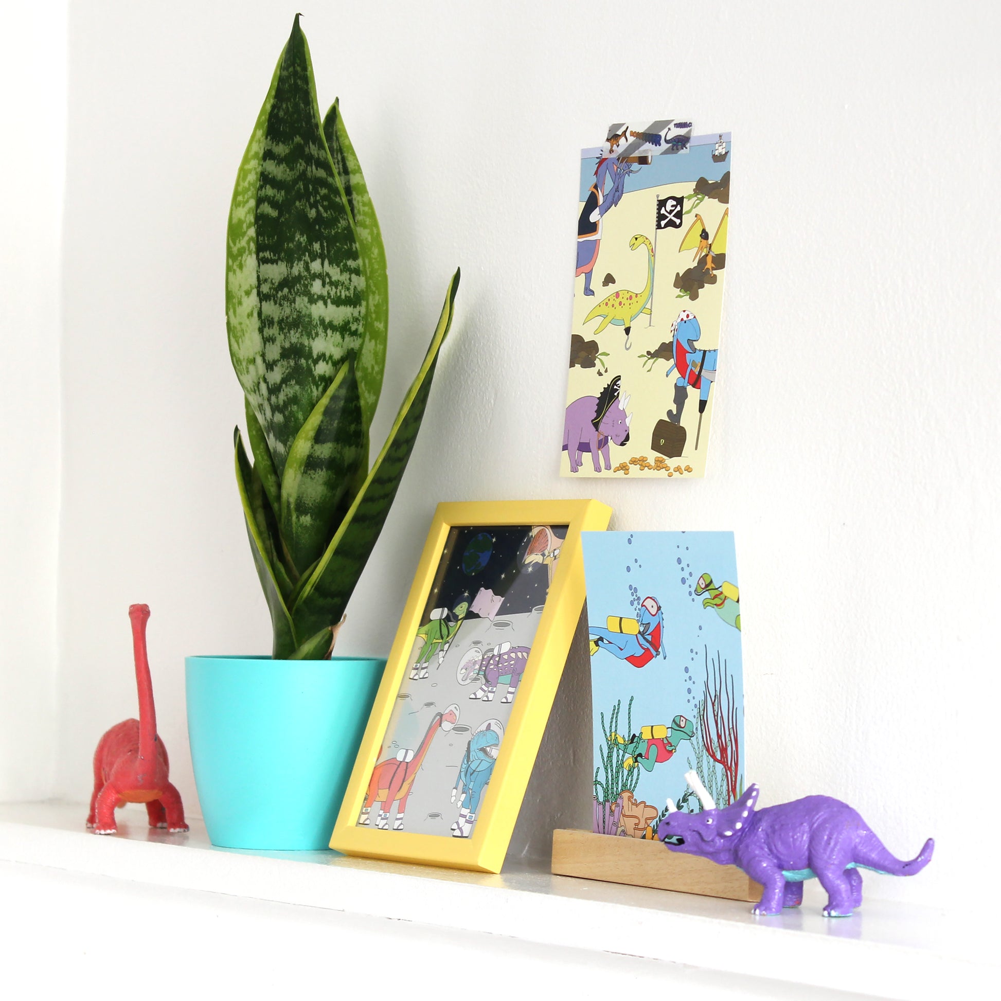 3 dinosaur postcards on a shelf with plants and dinosaur figurines 