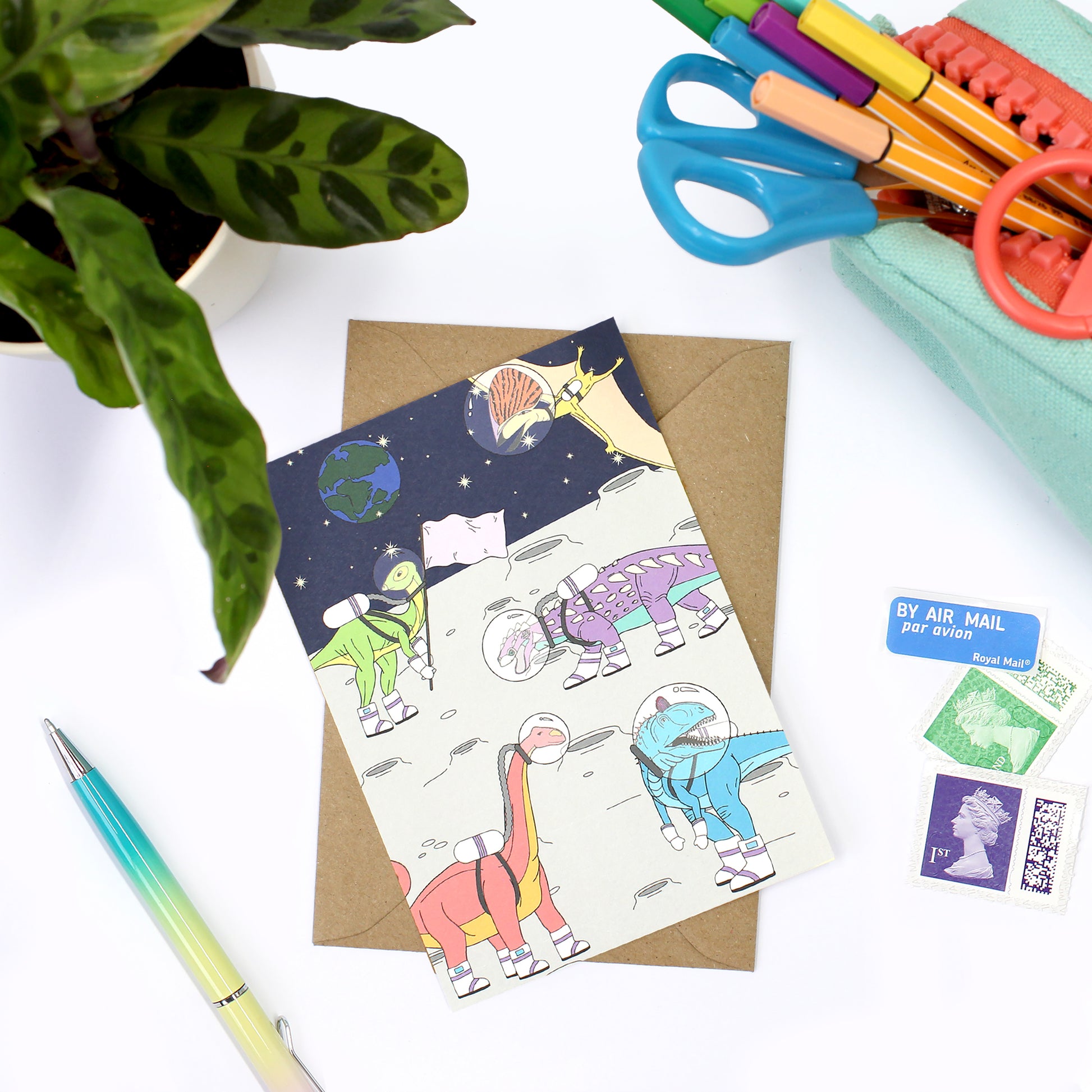 space dinosaur postcard on a desk with an envelope, stamps, pencil case and a plant