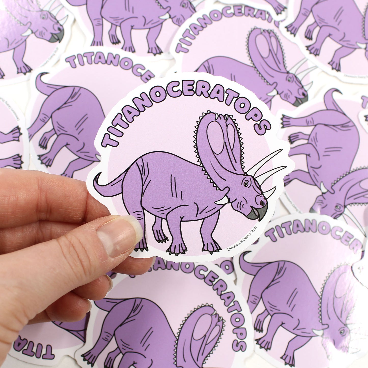 Titanoceratops dinosaur sticker held by a hand with scattered stickers behind it