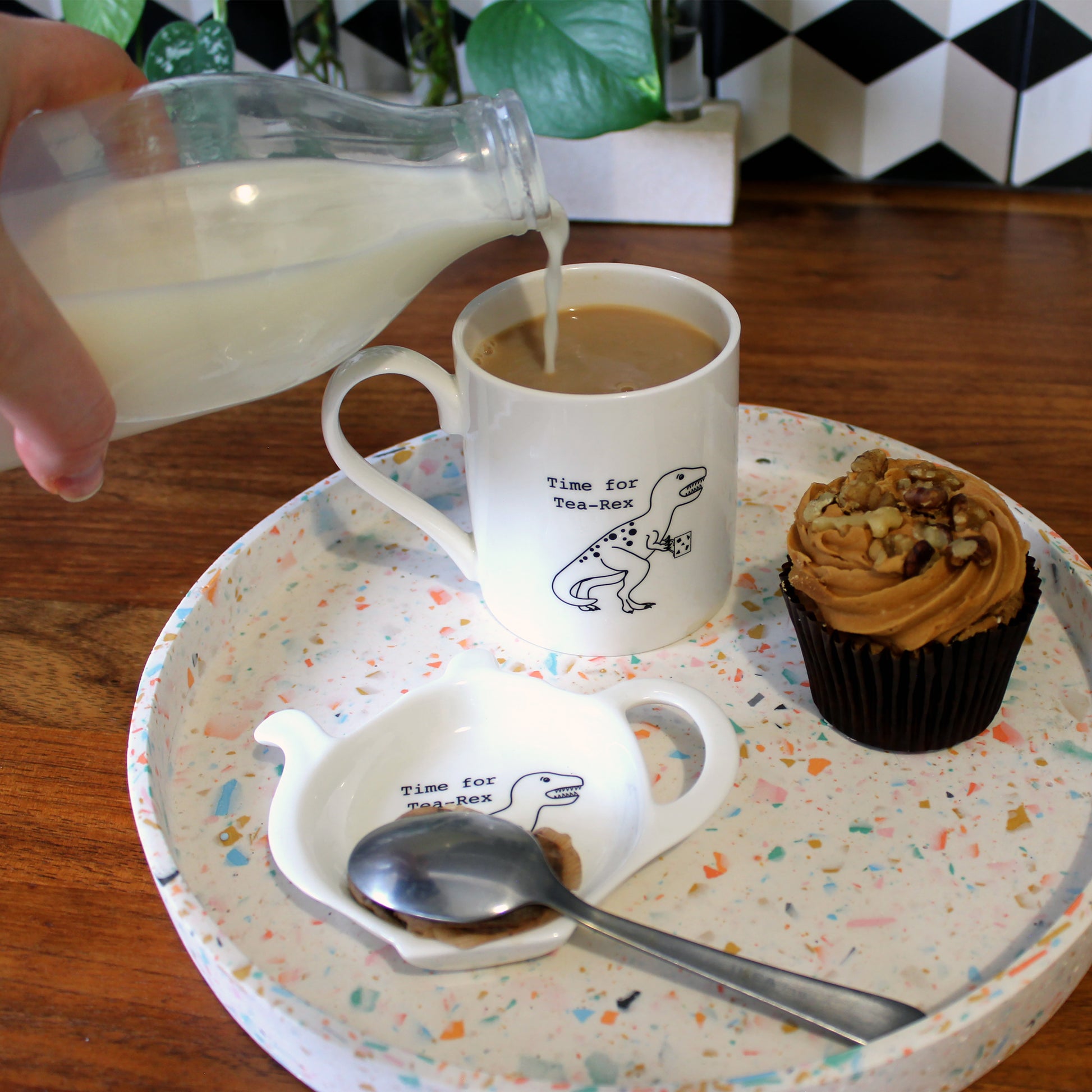 Time for Tea-Rex Mug and Teabag Tidy set on a terrazzo tray with a cupcake. A hand is pouring milk into the mug that is full of tea