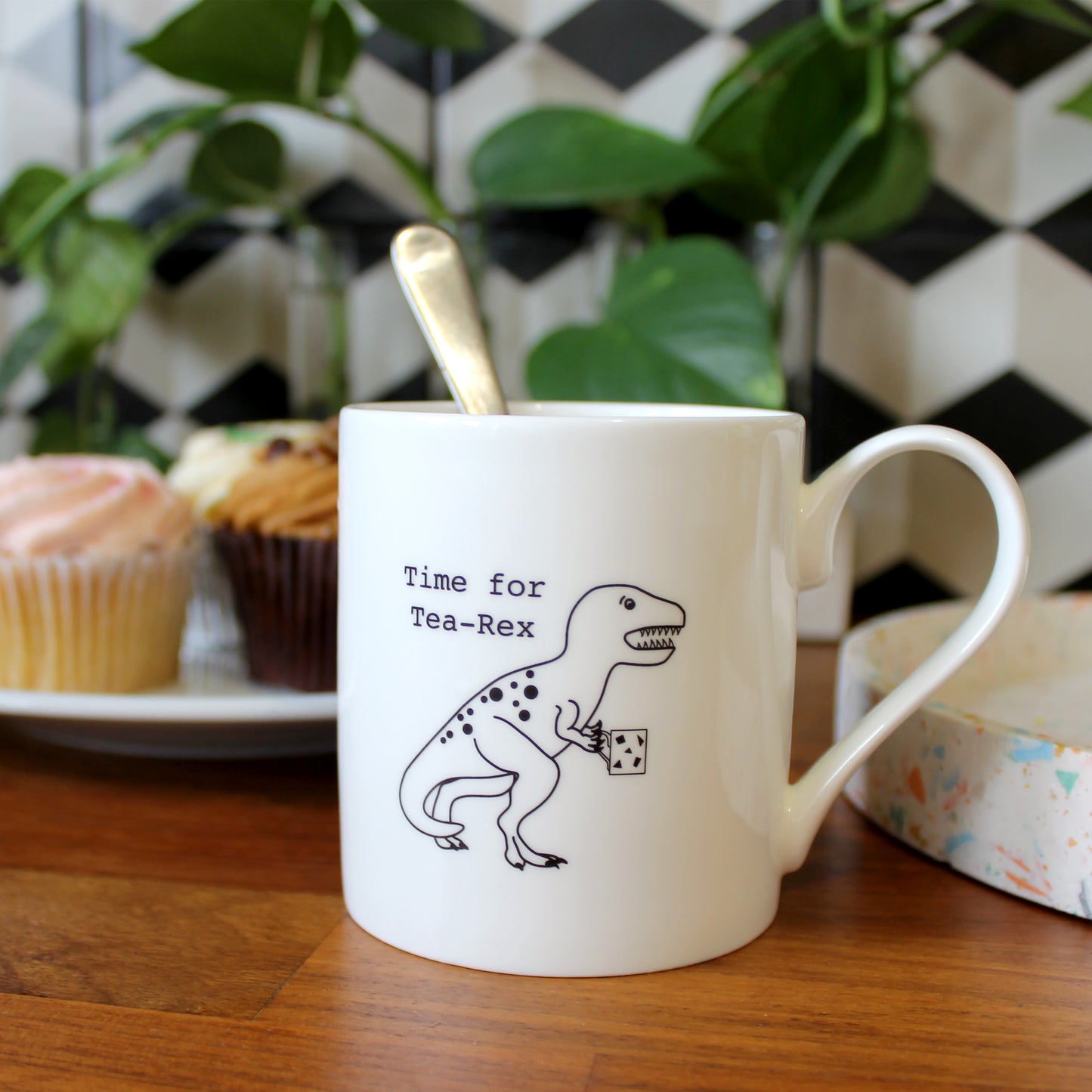 Time for Tea-Rex Mug in a kitchen with plants and cupcakes behind it