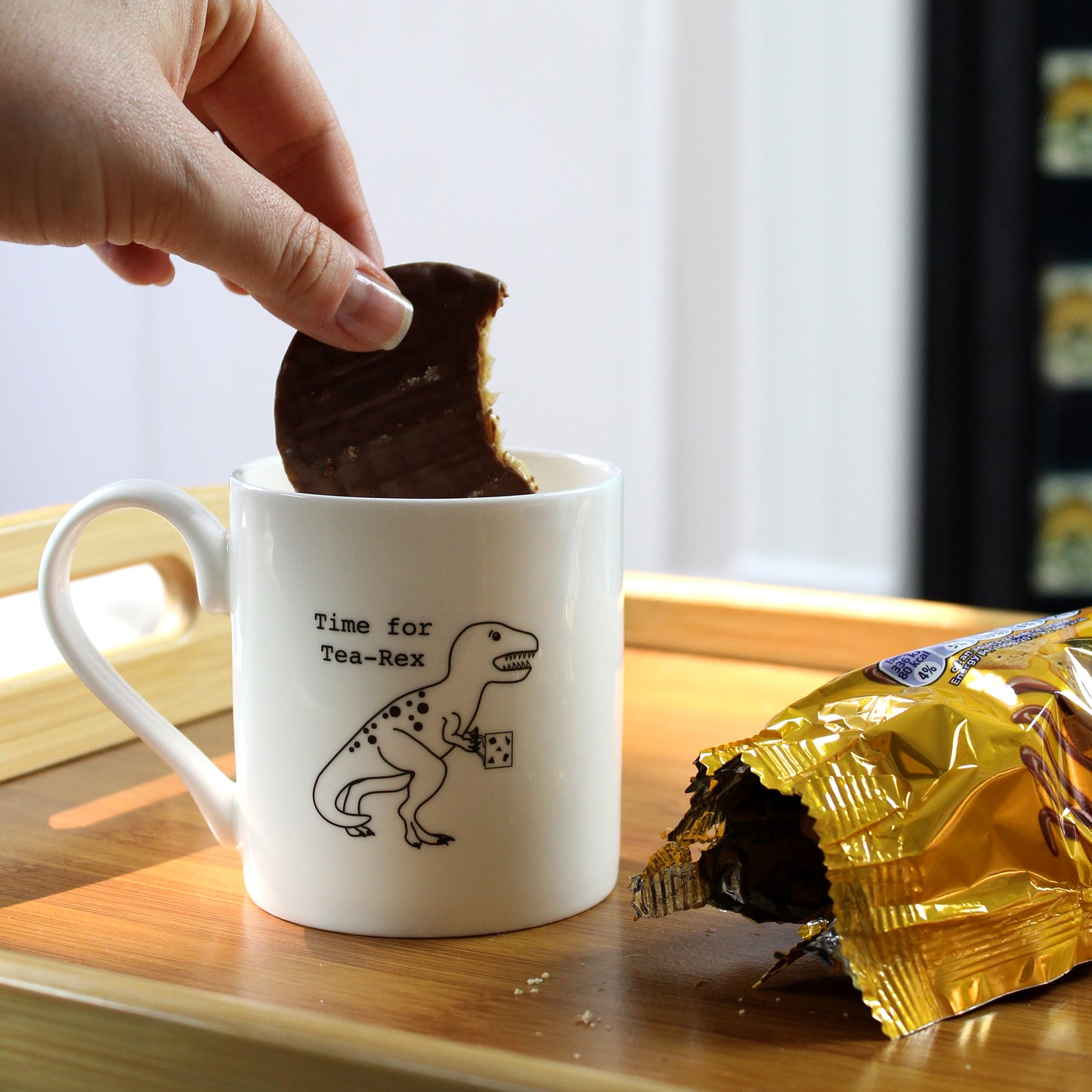 A hand dipping a partially eaten chocolate biscuit into a Time for Tea-Rex Mug