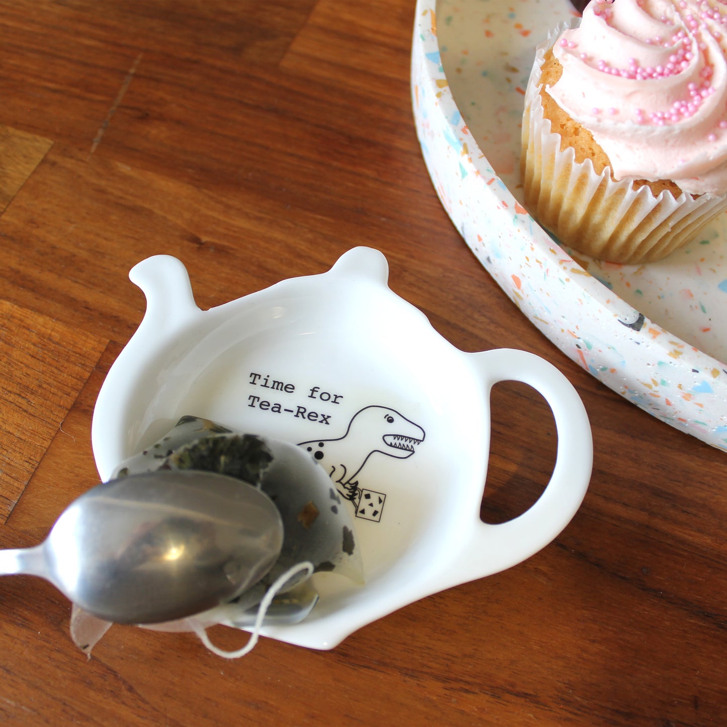 Time for Tea-Rex Teabag Tidy with a teabag and spoon resting on it. There is a pink cupcake on a terrazzo tray next to it